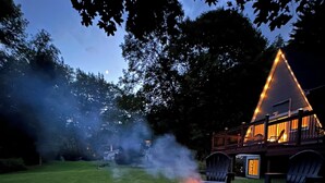 A fire pit with smoke, surrounded by stone tiles, in a grassy area with trees and a building with a triangular roof in the background.