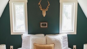 A bedroom with a large bed, two windows, a mounted deer head, and a plaid blanket.