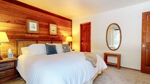 A bedroom with a wooden headboard, a bed with white bedding, a bedside table with a lamp, a mirror, and a door. - Mammoth Sierra Townhomes #38 3 Bedroom Condo (Mammoth Lakes)