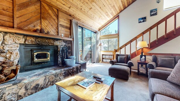 A cozy living room with a stone fireplace, wooden ceiling, and a staircase leading upstairs. - Mammoth Sierra Townhomes #38 3 Bedroom Condo (Mammoth Lakes)