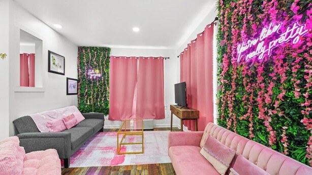 Un salon moderne avec un canapé gris, un canapé rose et une table basse en bois. On y trouve un téléviseur posé sur un meuble en bois et un néon fixé au mur.