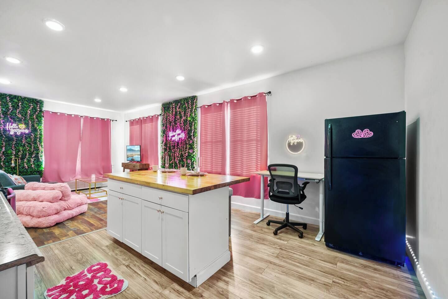 A modern kitchen with a central island, a refrigerator, and a dining area with pink curtains and a neon sign.