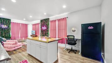 A modern kitchen with a central island, a refrigerator, and a dining area with pink curtains and a neon sign.
