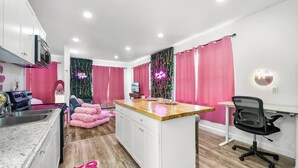 Une cuisine compacte dotée d’un îlot central, d’un coin repas avec des chaises roses et d’un espace de travail comprenant une chaise et un bureau.