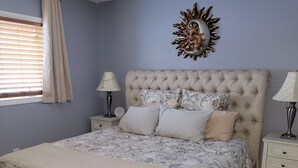 A bedroom with a tufted headboard, a floral-patterned bedspread, two bedside tables with lamps, and a decorative sunburst wall art piece.