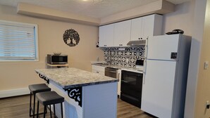A compact kitchen with white cabinets, a granite countertop, a microwave, a stove, and a refrigerator. There are two bar stools and a decorative wall piece.