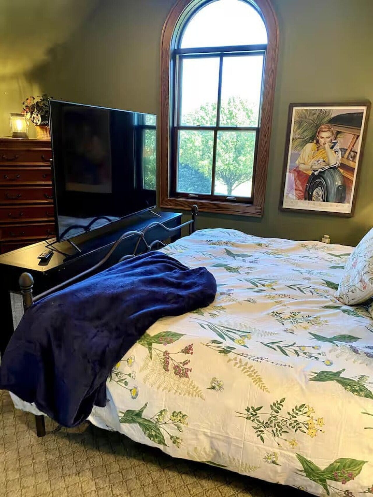 A bedroom with a bed, a TV, a dresser, and a window with a view of trees.
