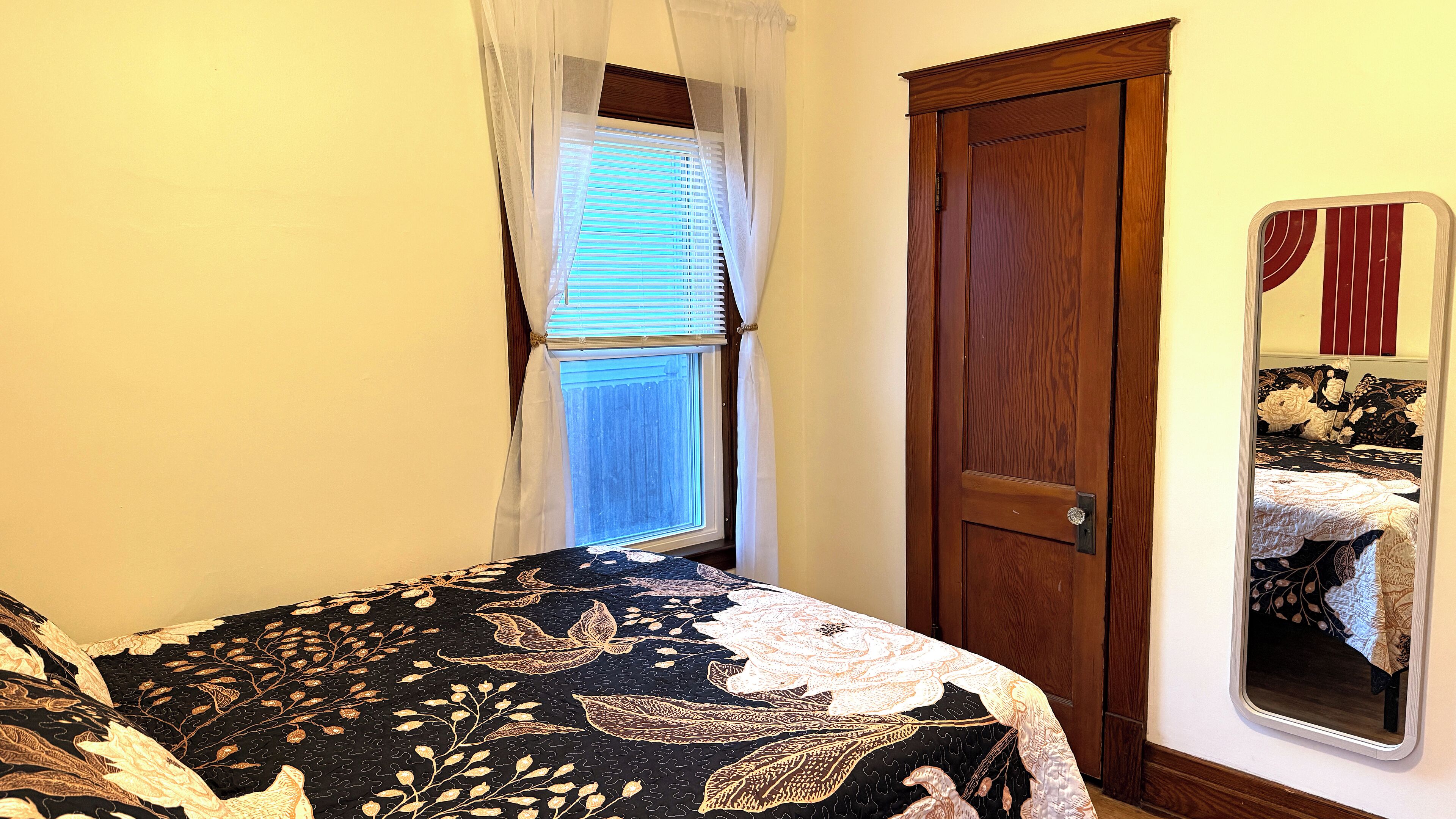 Un dormitorio con una cama, una ventana con persianas, una puerta de madera y un espejo en la pared.