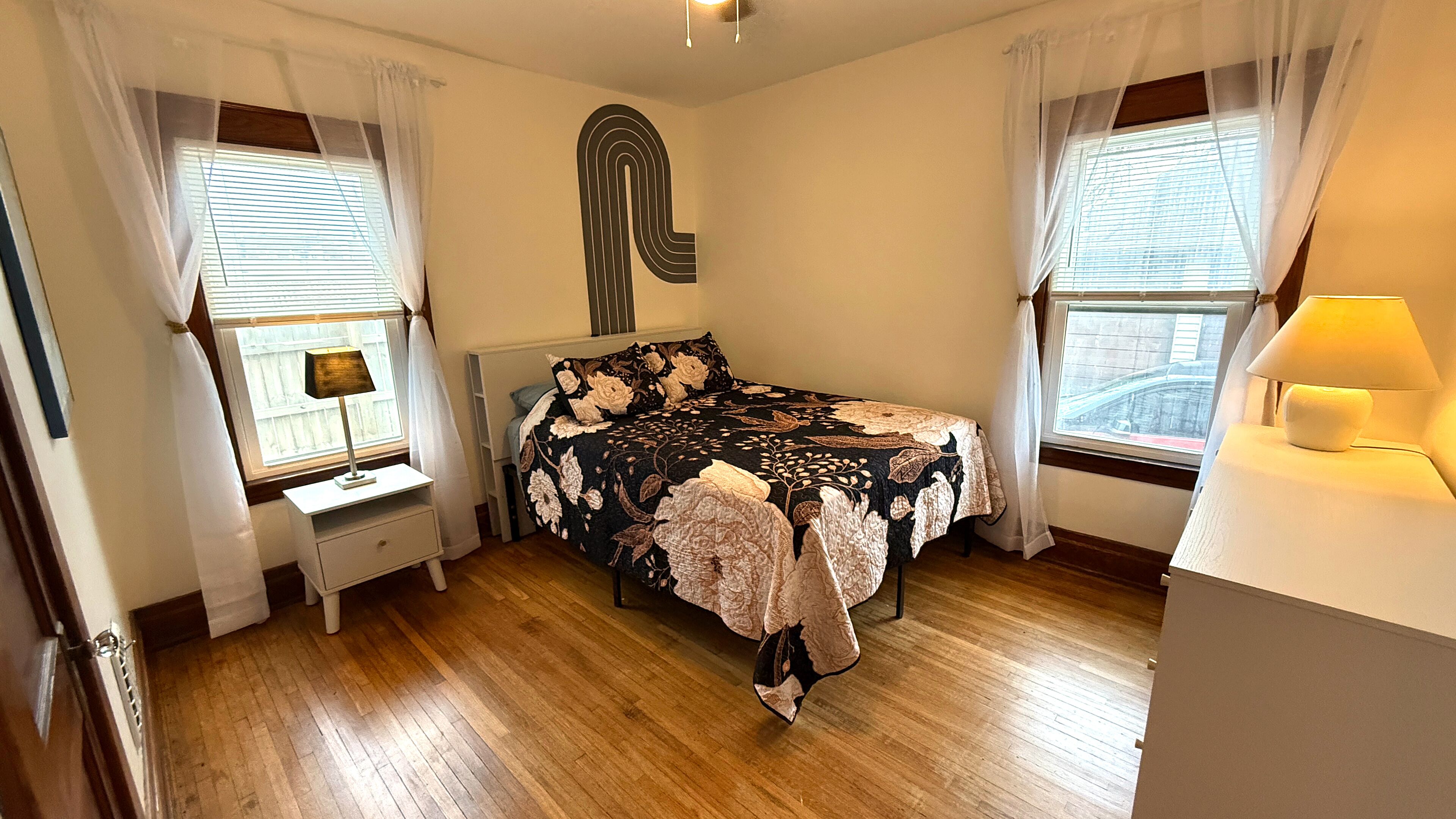 Un dormitorio con cama, mesita de noche, lámpara y ventana con cortinas.