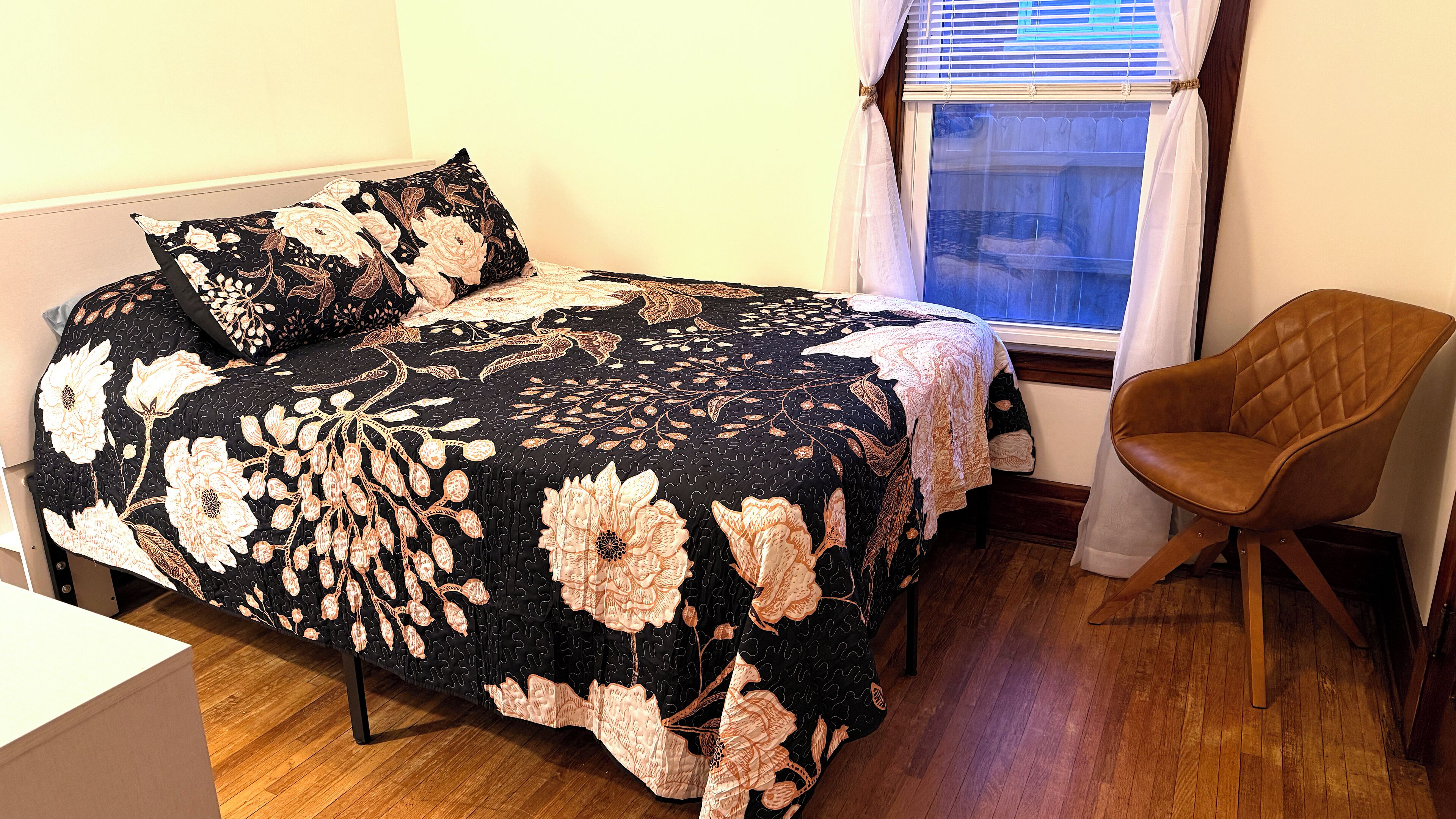 A bedroom with a bed covered in a floral-patterned bedspread, a wooden chair, and a window with blinds.