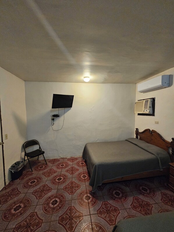 A small, single-bed room with a patterned floor, a television mounted on the wall, and an air conditioning unit. - Hotel Chalet (Matamoros)