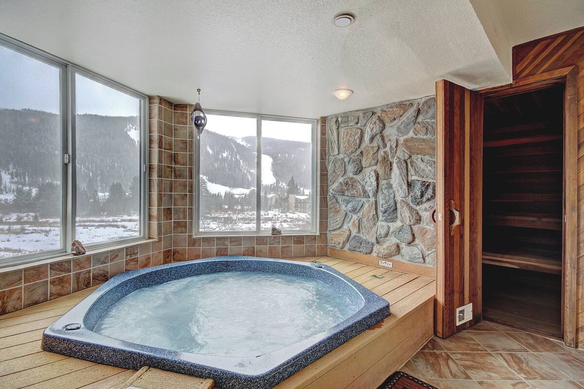 Un jacuzzi con pared de piedra y piso de madera, ventanas amplias con vista a una montaña nevada y una puerta de madera.