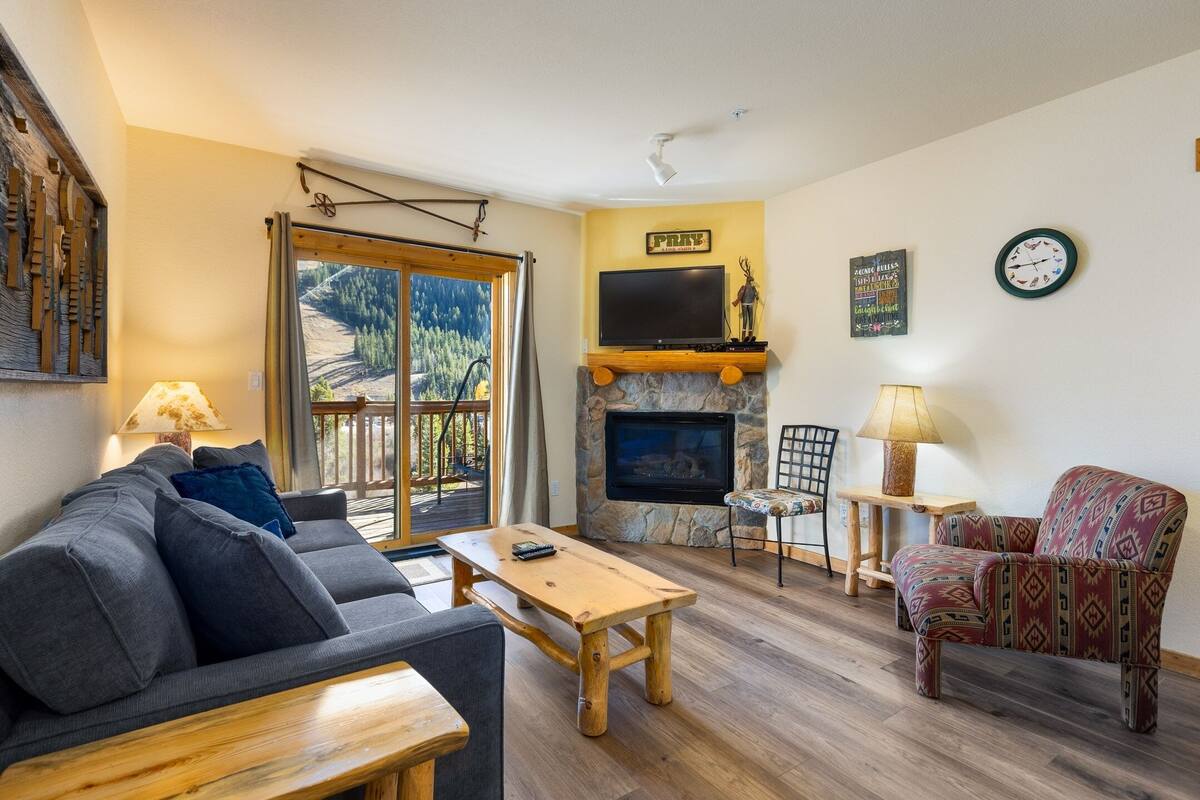 Una sala de estar con un sofá, una mesa de centro de madera, un sillón estampado, una chimenea de piedra, un televisor montado sobre la chimenea y un ventanal con vistas a árboles y montañas.