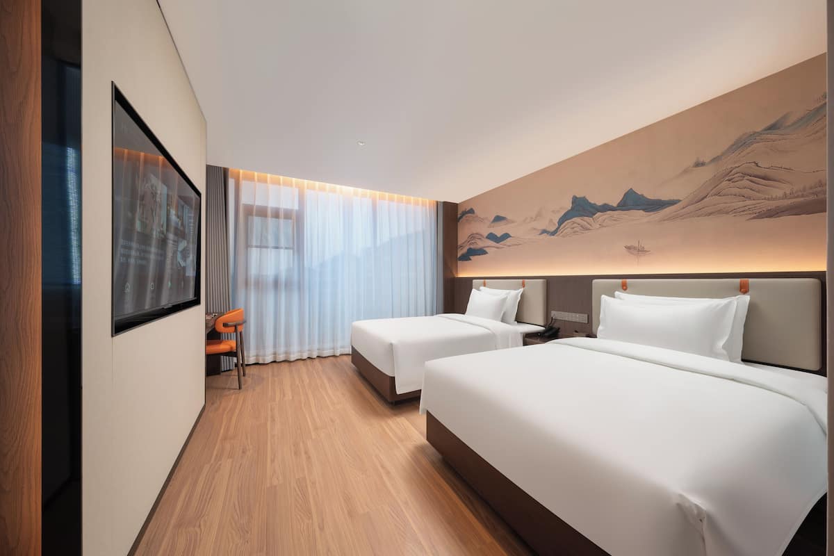 Habitación de hotel con dos camas, piso de madera, un cuadro grande en la pared y una ventana con cortinas de gasa.