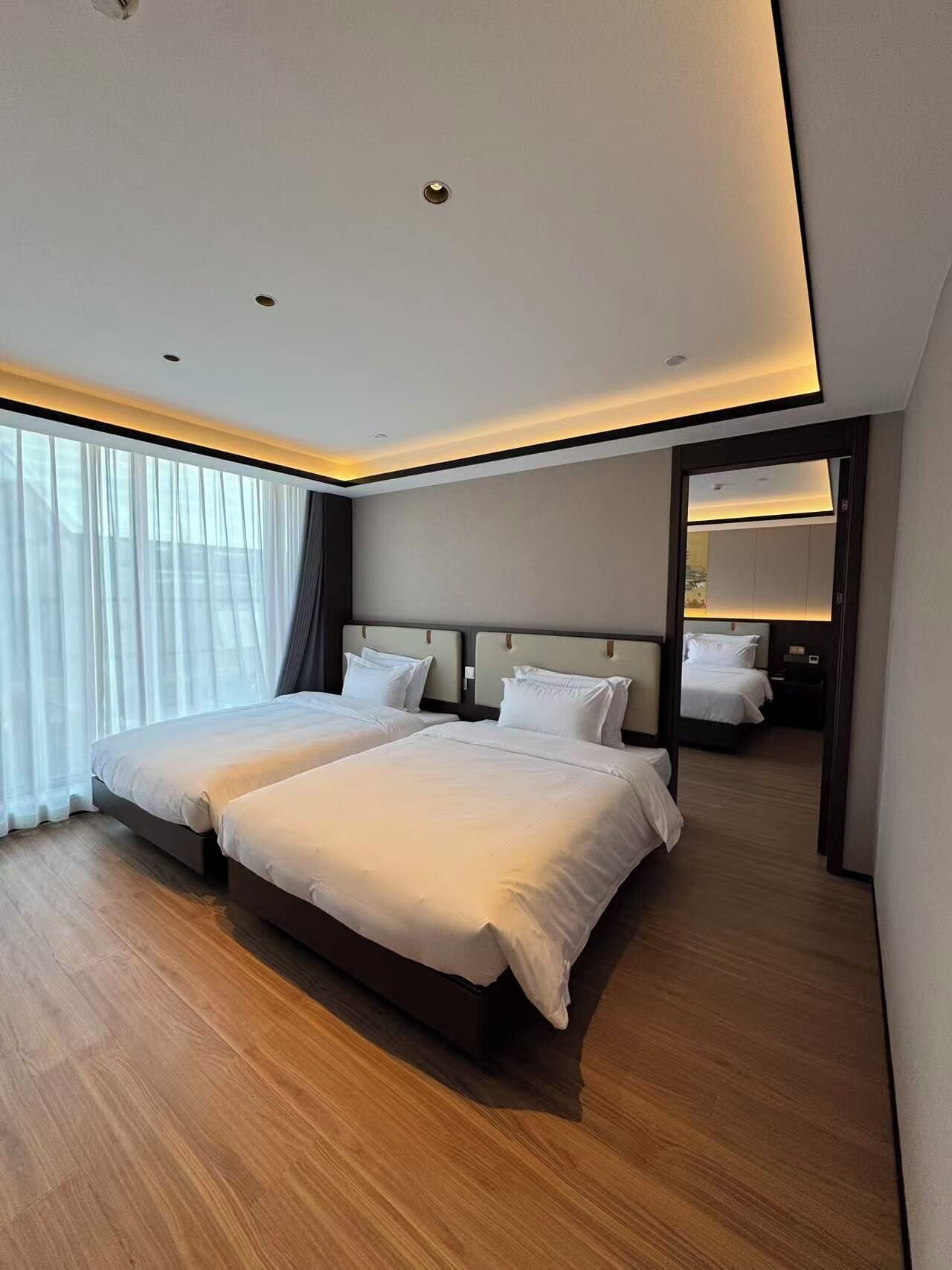 A modern hotel room with two beds, a large window with sheer curtains, and a mirror reflecting the room.