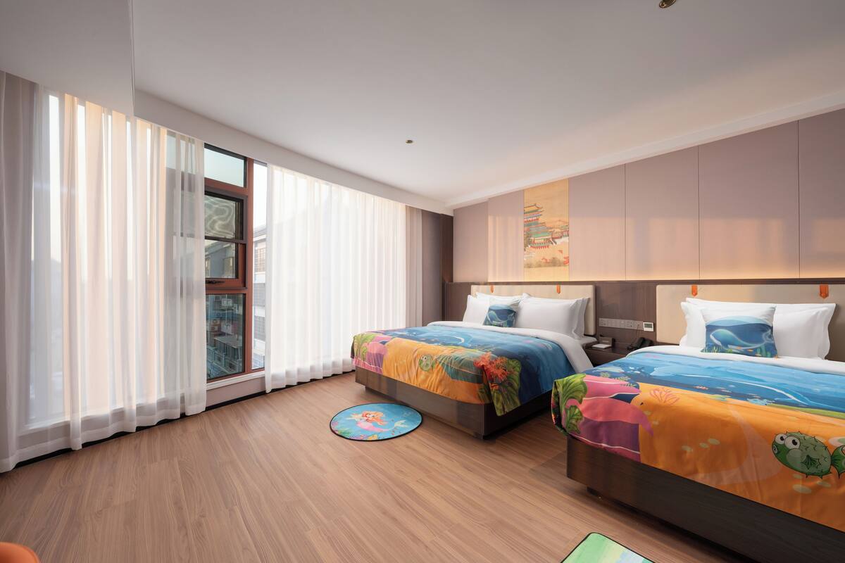 Una habitación de hotel moderna con dos camas, ventanas amplias con cortinas translúcidas, piso de madera y una colcha colorida.