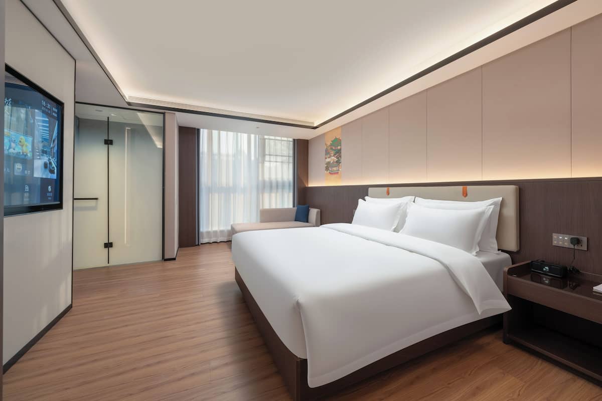 Una habitación de hotel moderna con una cama grande, una mesita de noche con lámpara, un televisor montado en la pared y una puerta corredera que da a un balcón.