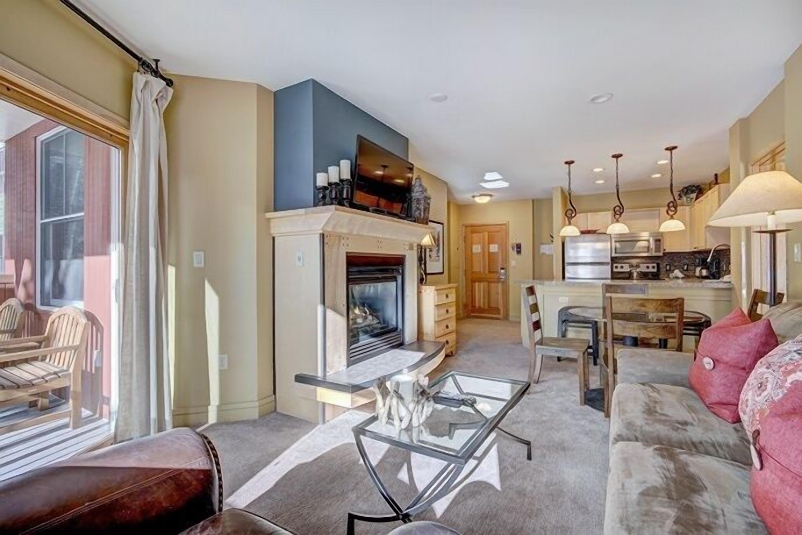 A living room with a fireplace, a glass coffee table, a sofa, and a kitchen area in the background.