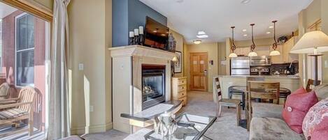 A living room with a fireplace, a glass coffee table, a sofa, and a kitchen area in the background.