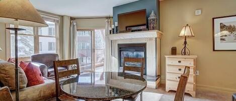 A living room with a glass-top table, wooden chairs, a sofa, a fireplace, and a television on a stand.