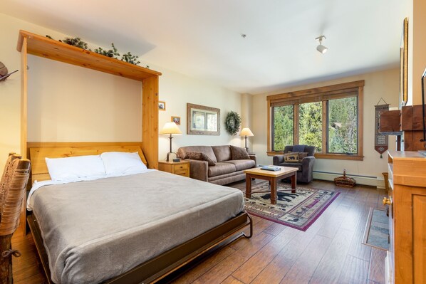 A bedroom with a large bed, a wooden headboard, a nightstand, a sofa, a coffee table, and a window with a view of trees.