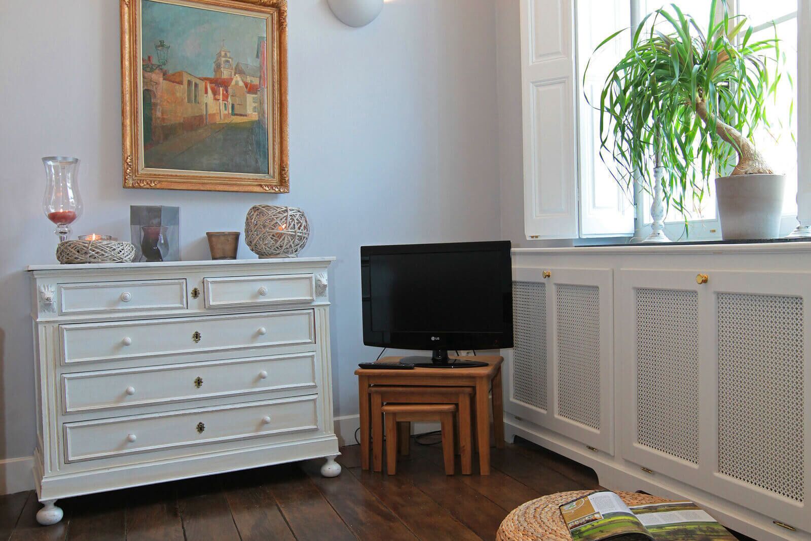 A room with a white dresser, a television on a wooden stand, a potted plant, and a painting on the wall.