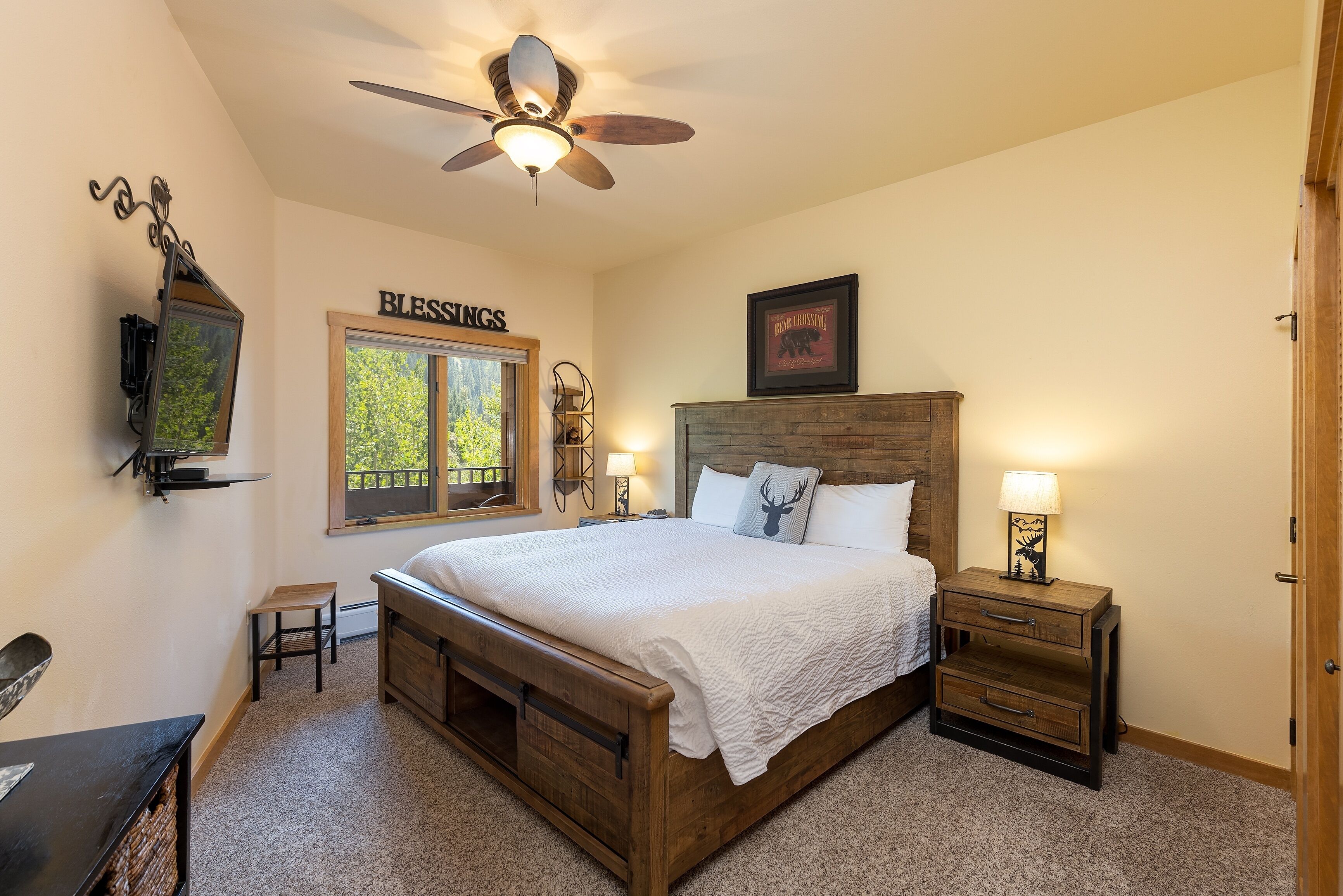 A bedroom with a wooden bed, a nightstand, a ceiling fan, a window with a view, and a wall-mounted shelf.
