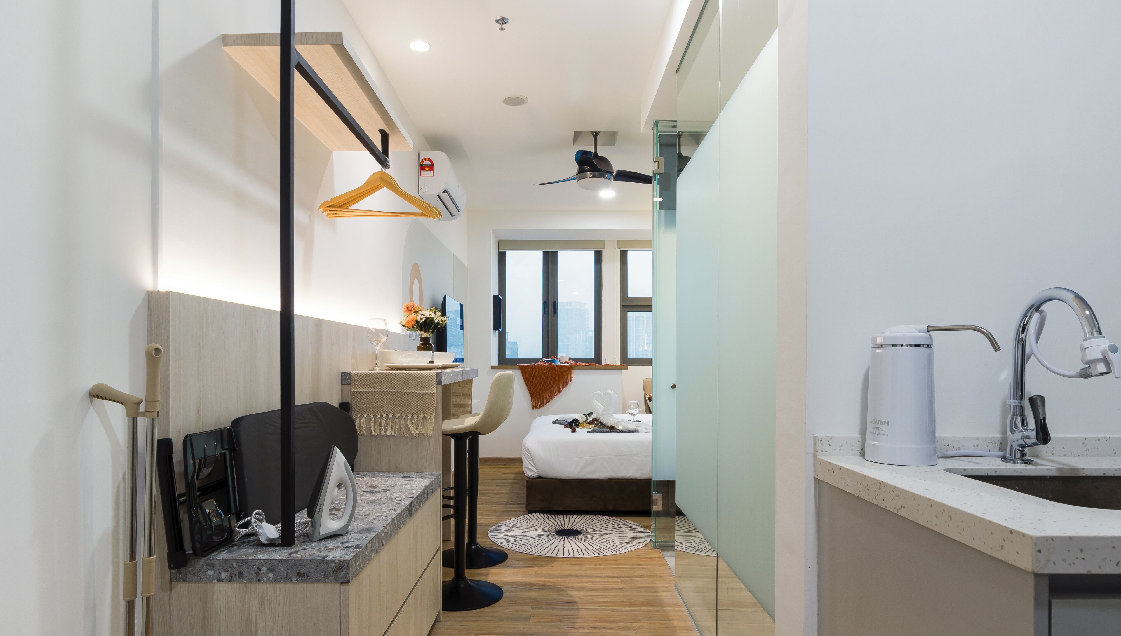A modern hotel room with a bed, a small sitting area with a chair and table, a kitchenette, and a bathroom with a sink and shower.