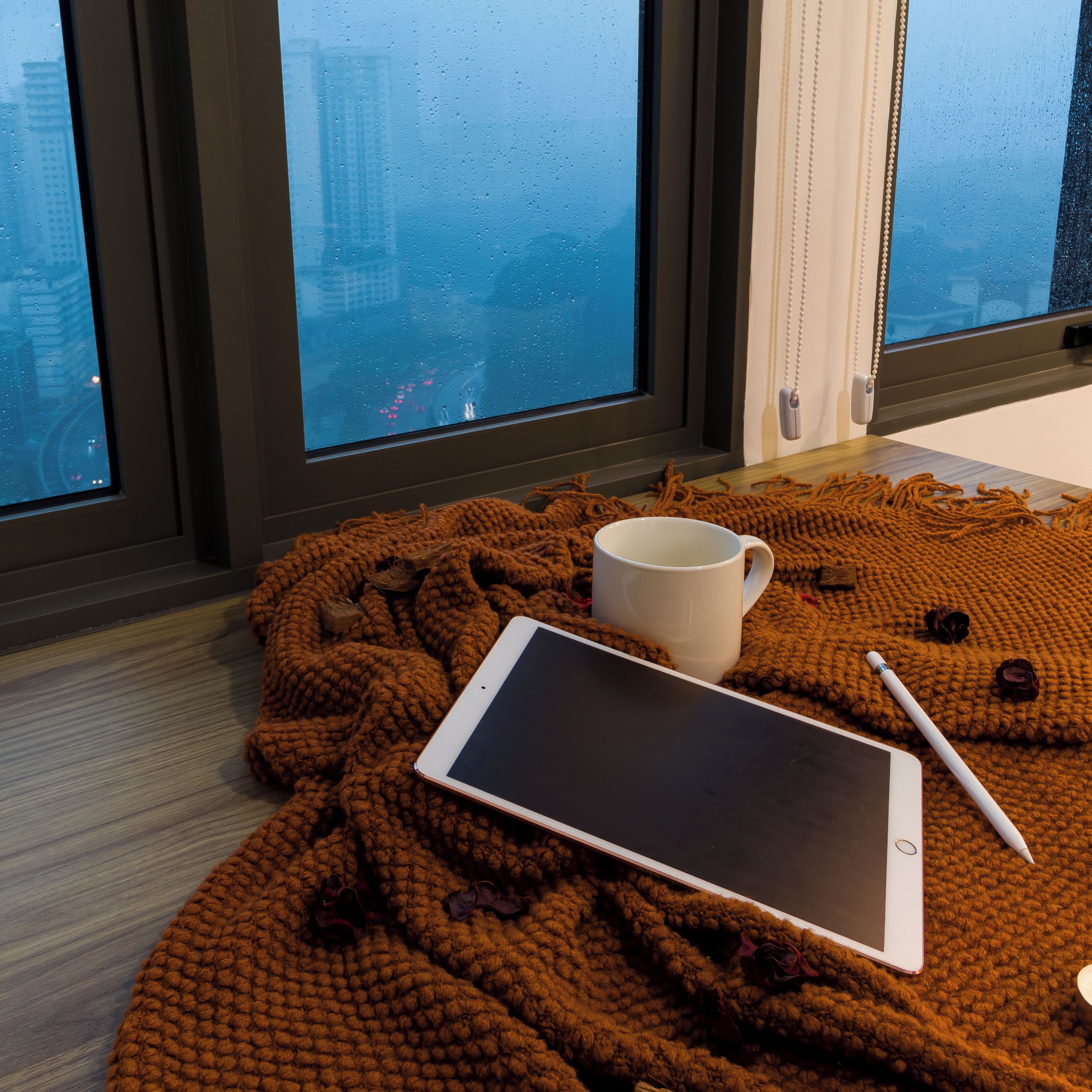 Un espace intérieur chaleureux et accueillant, avec une tablette, une tasse et un stylo posés sur une couverture tricotée, et une fenêtre donnant sur un paysage urbain.