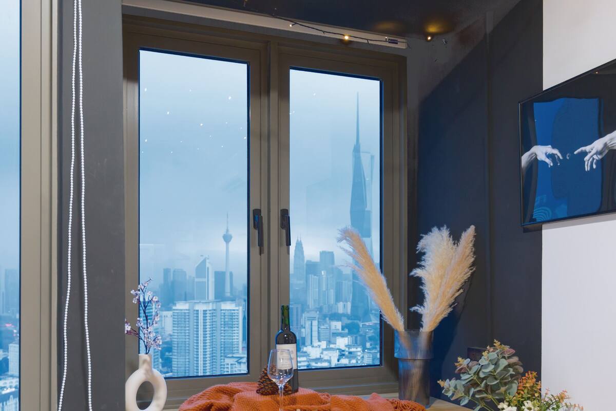 A room with a large window overlooking a cityscape, a wooden table with a wine glass, a vase with pampas grass, and a framed artwork on the wall.