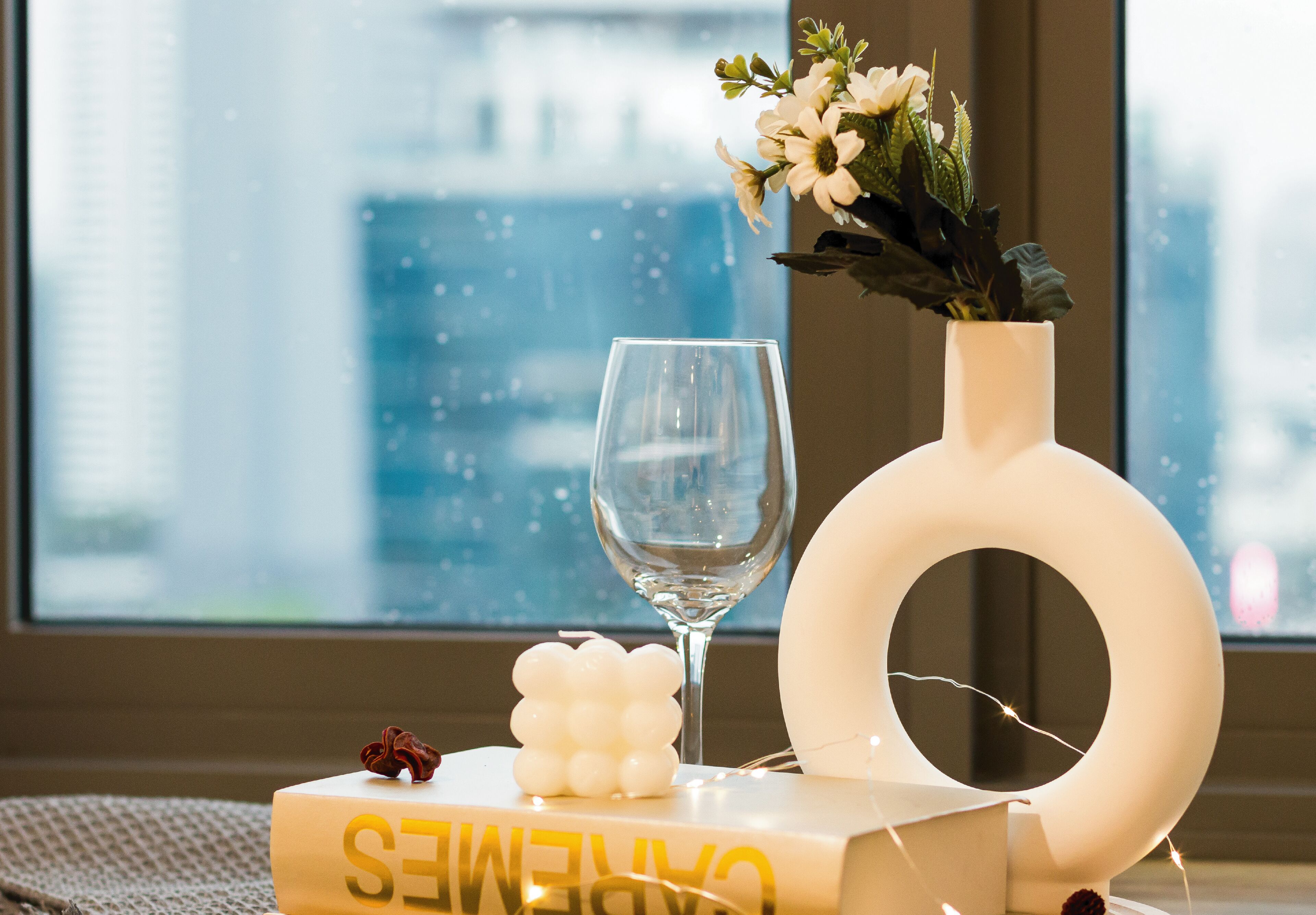 A room with a large window, a vase with flowers, a wine glass, and a decorative object on a table.