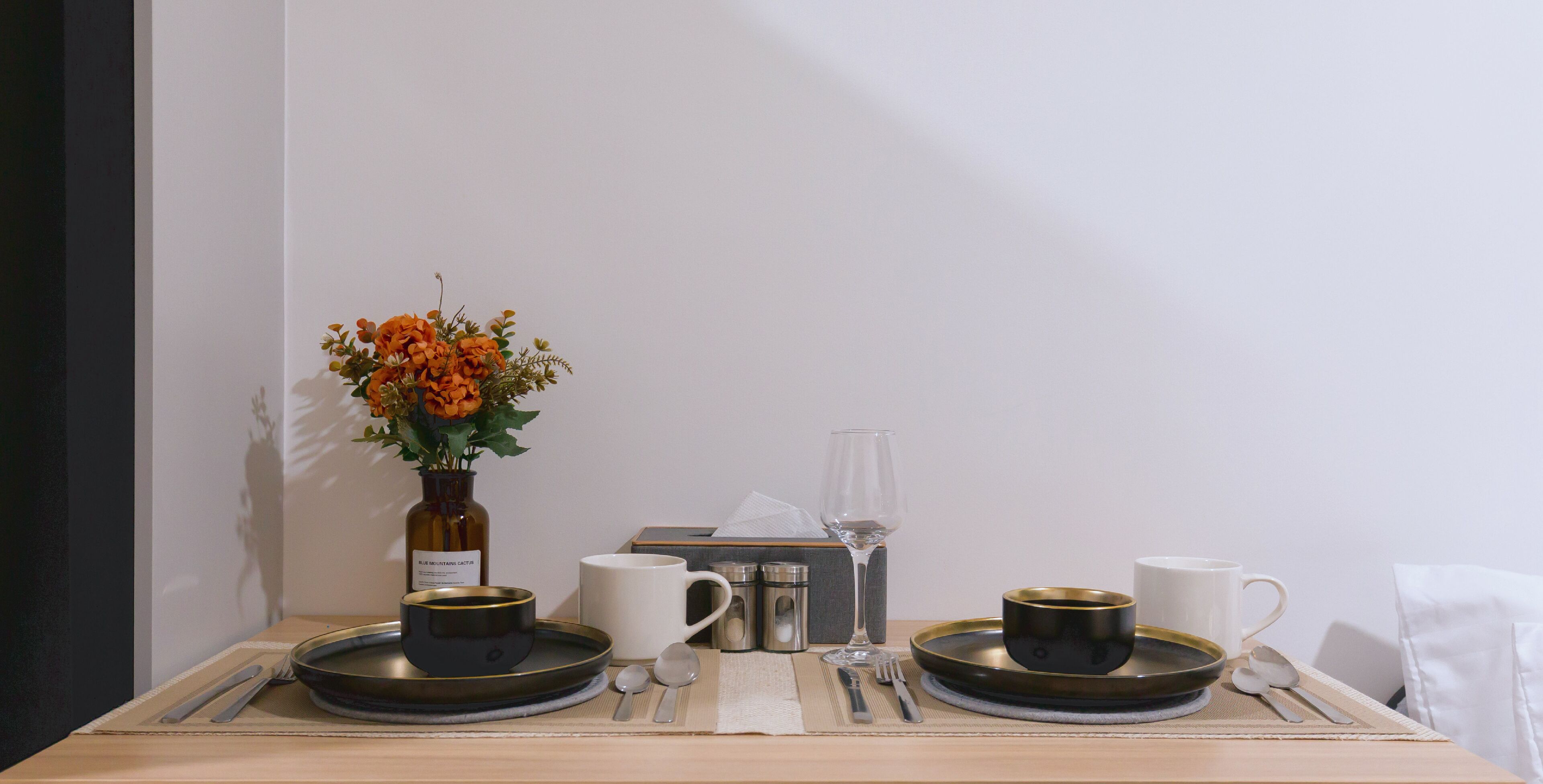 A neatly set dining table with a vase of flowers, two black cups, a white cup, and a glass.