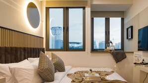 A modern hotel room with a large bed, two windows with city views, a round mirror, and a bedside table with a book and a vase. - The Riv Riveria City KL Sentral (Kuala Lumpur)