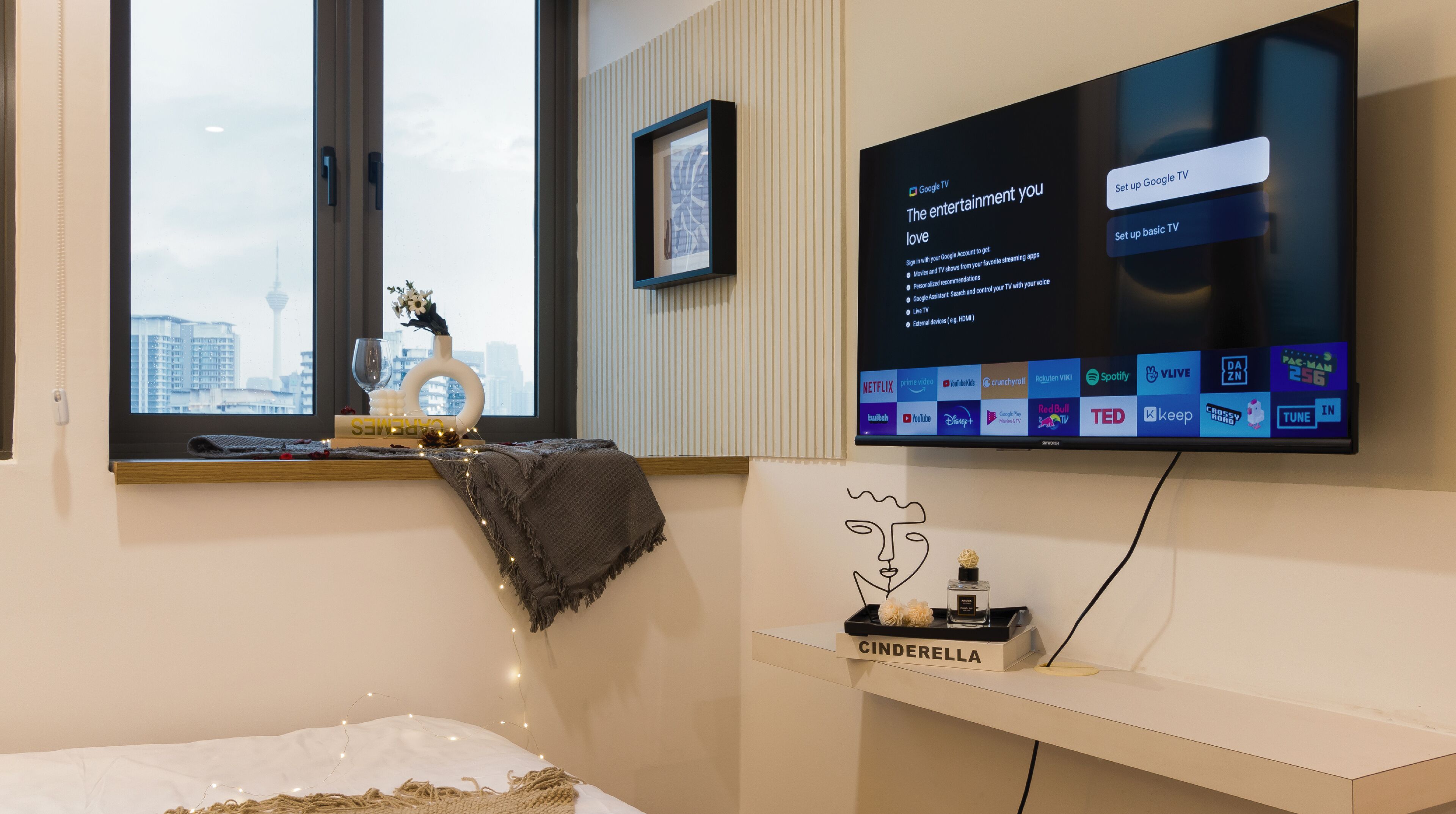 A modern hotel room with a large flat-screen TV mounted on the wall, a window with a view of buildings, a bedside table with a decorative item, and a bedside lamp.