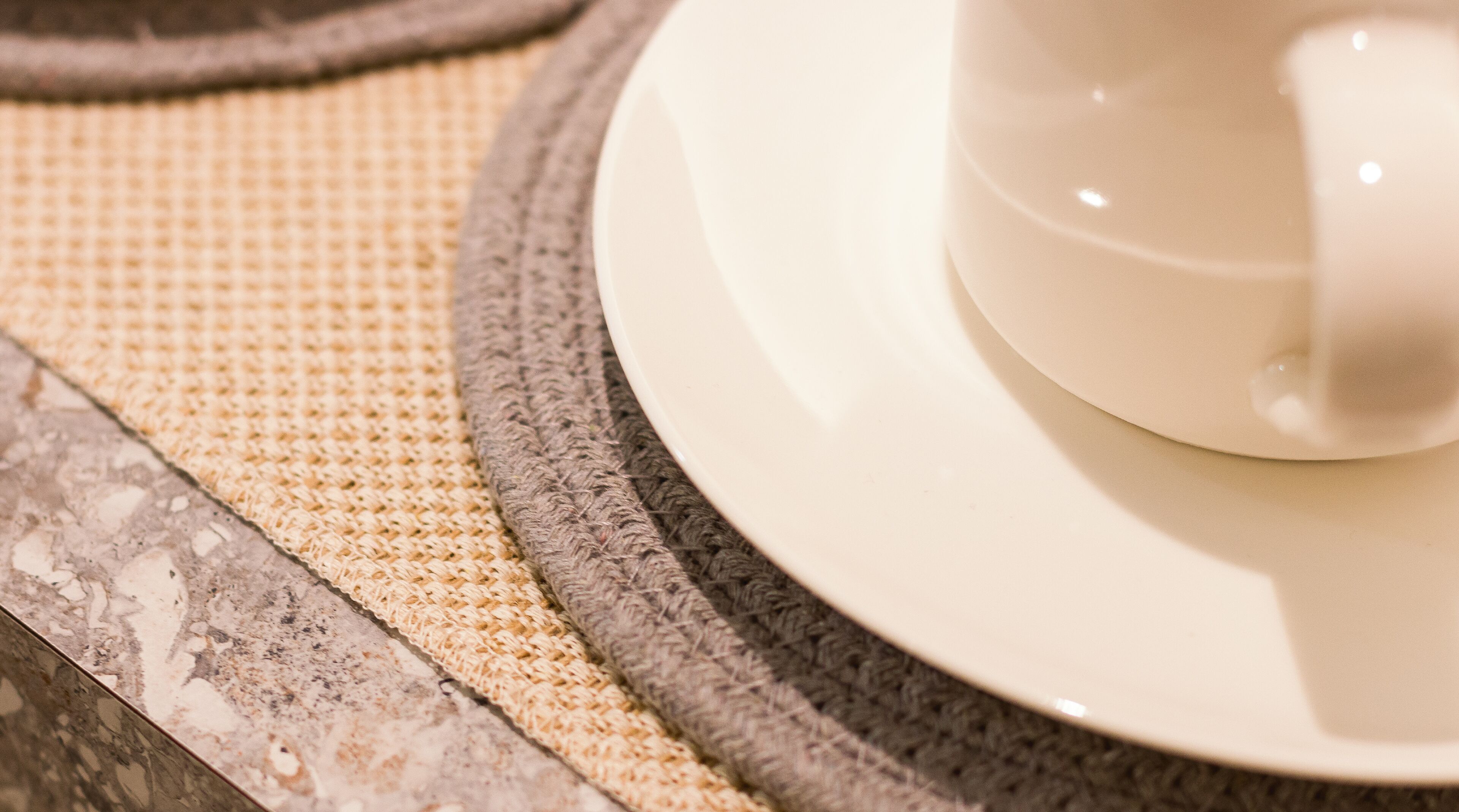 A close-up of a white ceramic cup on a matching saucer, placed on a textured woven mat, with a marble countertop in the background.