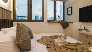 Habitación de hotel moderna con una cama grande, un ventanal con vistas a la ciudad, un televisor montado y una estantería con un adorno.