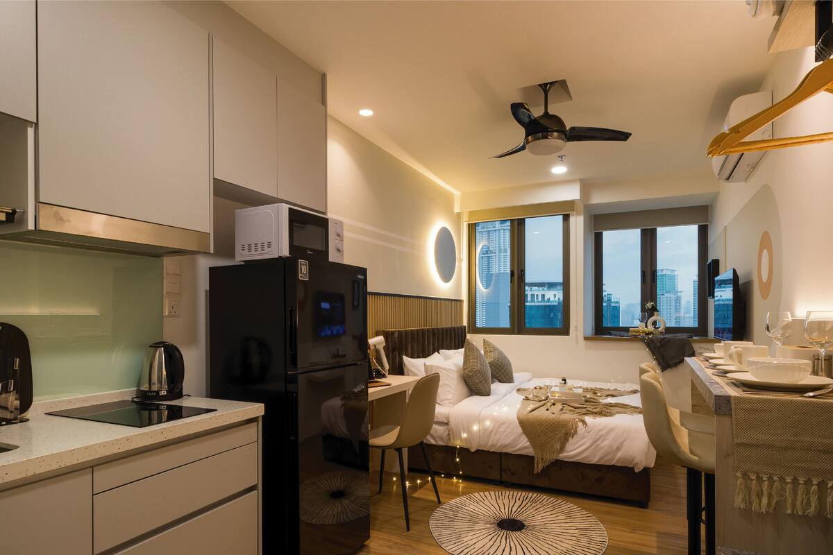 A compact hotel room with a kitchenette, a bed with a view of the city, a dining area, and a ceiling fan.
