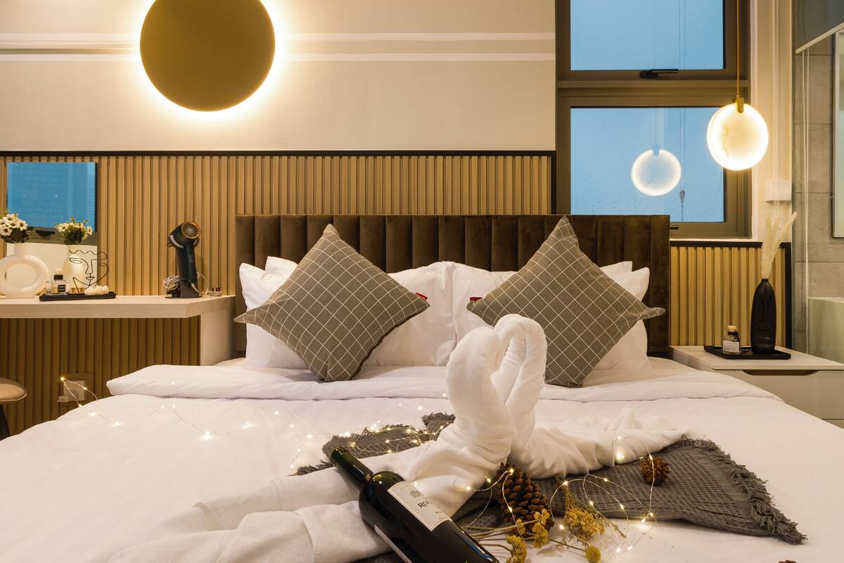 A hotel room with a large bed, two pillows, a bedside table with a lamp, a wall-mounted light, and a window with a view.