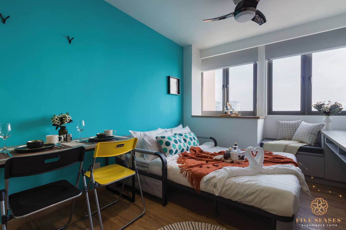 A modern hotel room with a teal accent wall, a bed with a white and orange blanket, a desk with a chair, and a window with a city view.