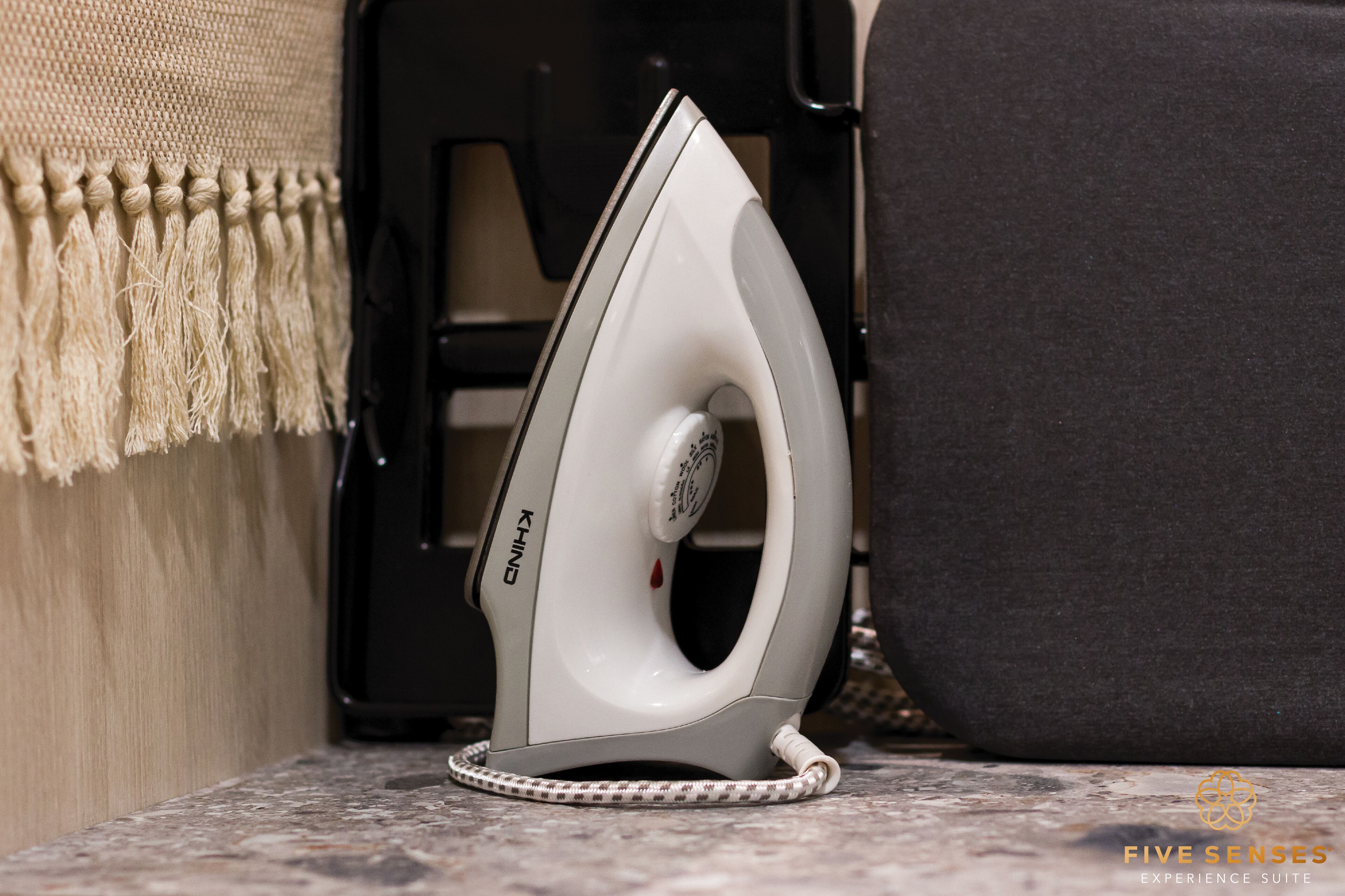 A white iron with a cord, placed on a stone surface near a black chair and a beige wall with a fringe.