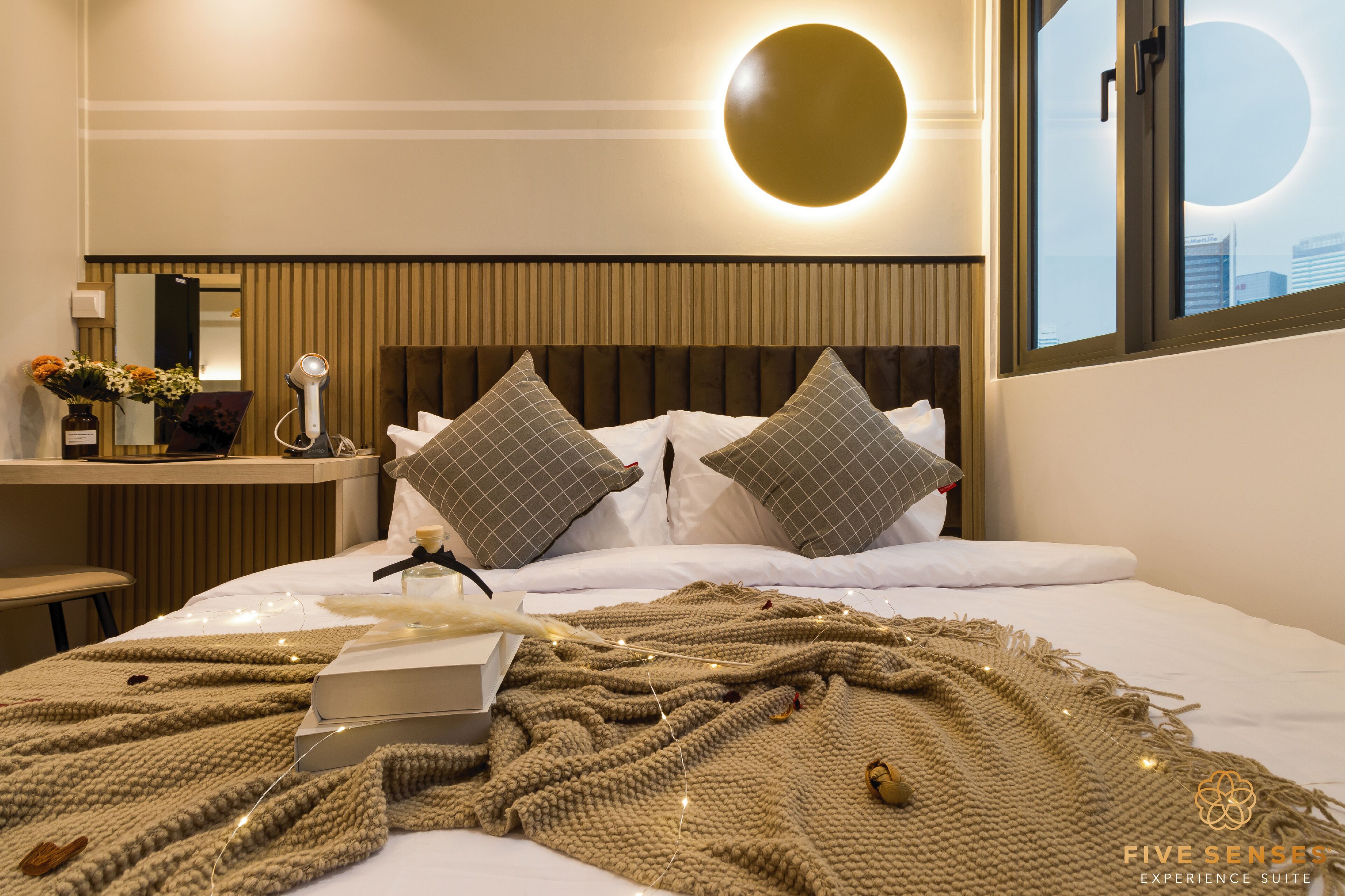 Een net opgemaakt bed met een geruit kussen, een nachtkastje met een lamp, en een wandlamp.