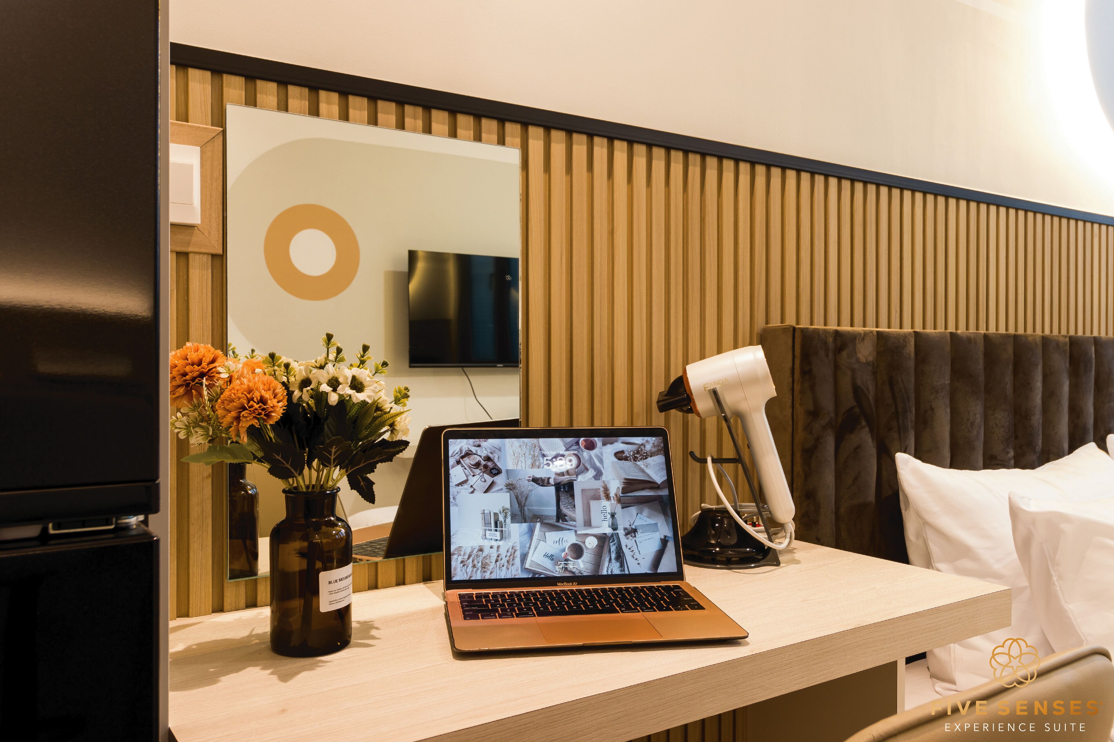 A hotel room with a wooden panel wall, a flat-screen TV mounted above a desk, a laptop, a hairdryer, and a vase with flowers.
