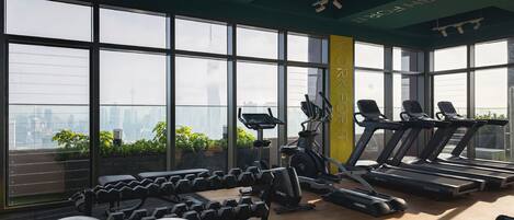 A gym with treadmills, dumbbells, and exercise balls, large windows, and a view of a cityscape.