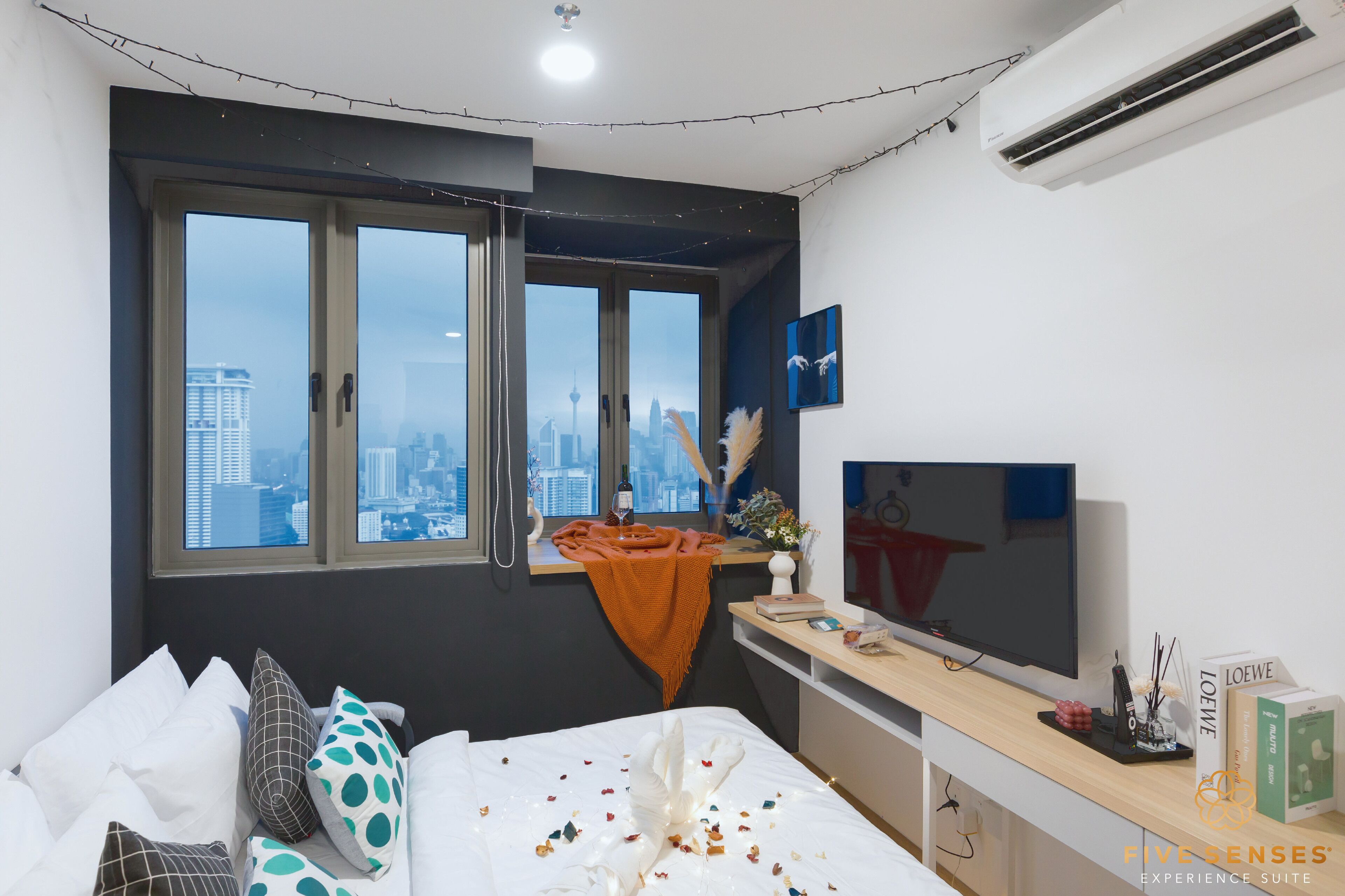 Een moderne hotelkamer met uitzicht op de stad, een bed met gekleurde kussens, een televisie en een klein bureau met een vaas en boeken.