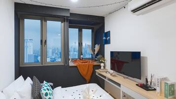 Een moderne hotelkamer met uitzicht op de stad, een bed met gekleurde kussens, een televisie en een klein bureau met een vaas en boeken.