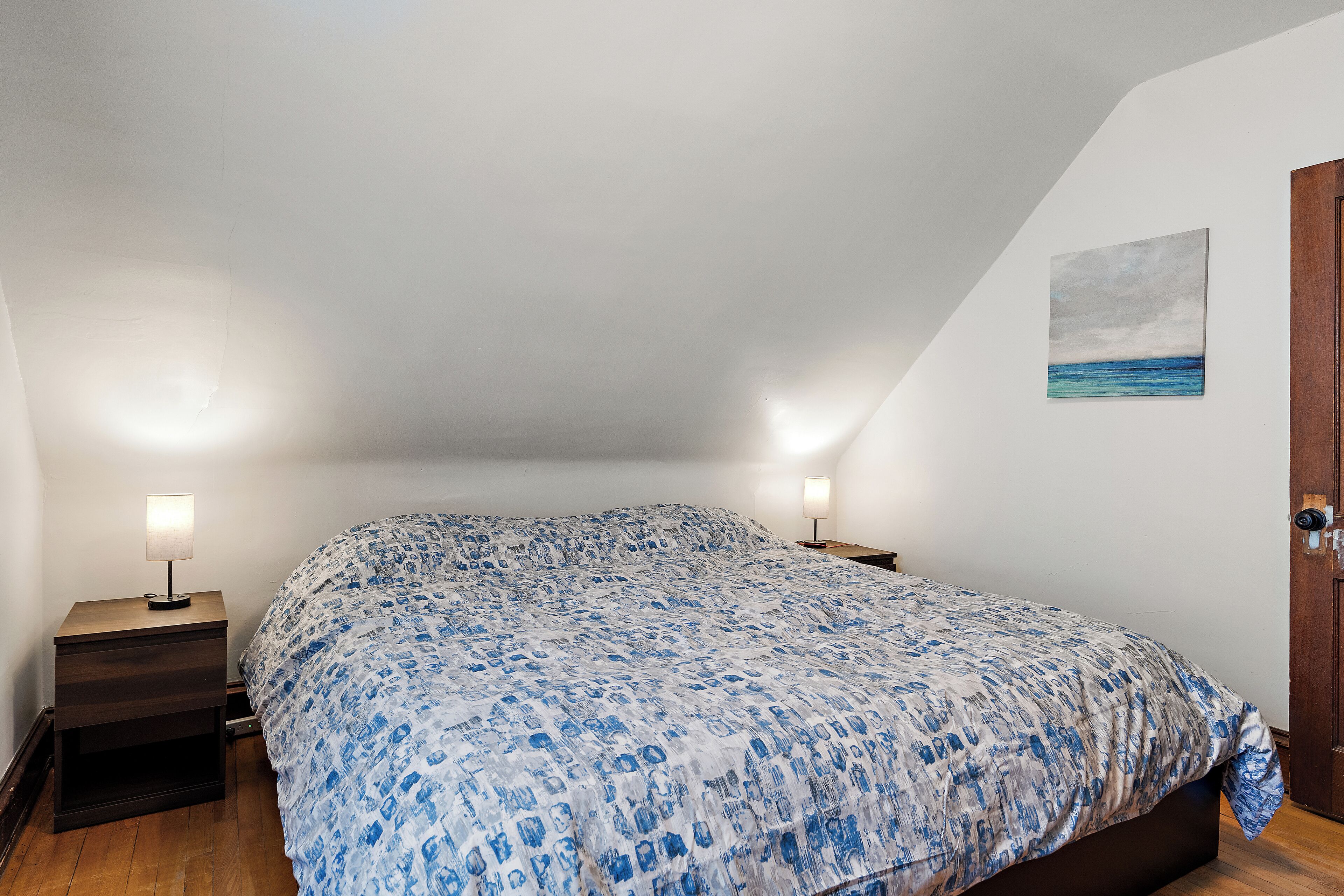 Quarto com uma cama grande, duas mesas de cabeceira com abajures, uma porta de madeira e um quadro na parede.