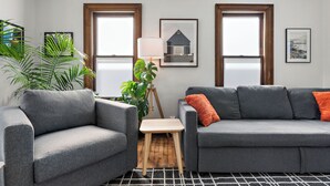 A living room with two grey sofas, a small wooden table, and a floor lamp. There are two windows with wooden frames and a view of the outside. - NordEast Escape/Hot Tub, Bocce court, Fooseball, Patio, Awesome location (Minneapolis)