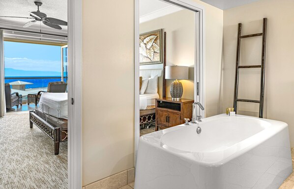 Un baño moderno con una bañera grande, un armario de madera y un espejo que refleja un dormitorio con cama y vista al mar.