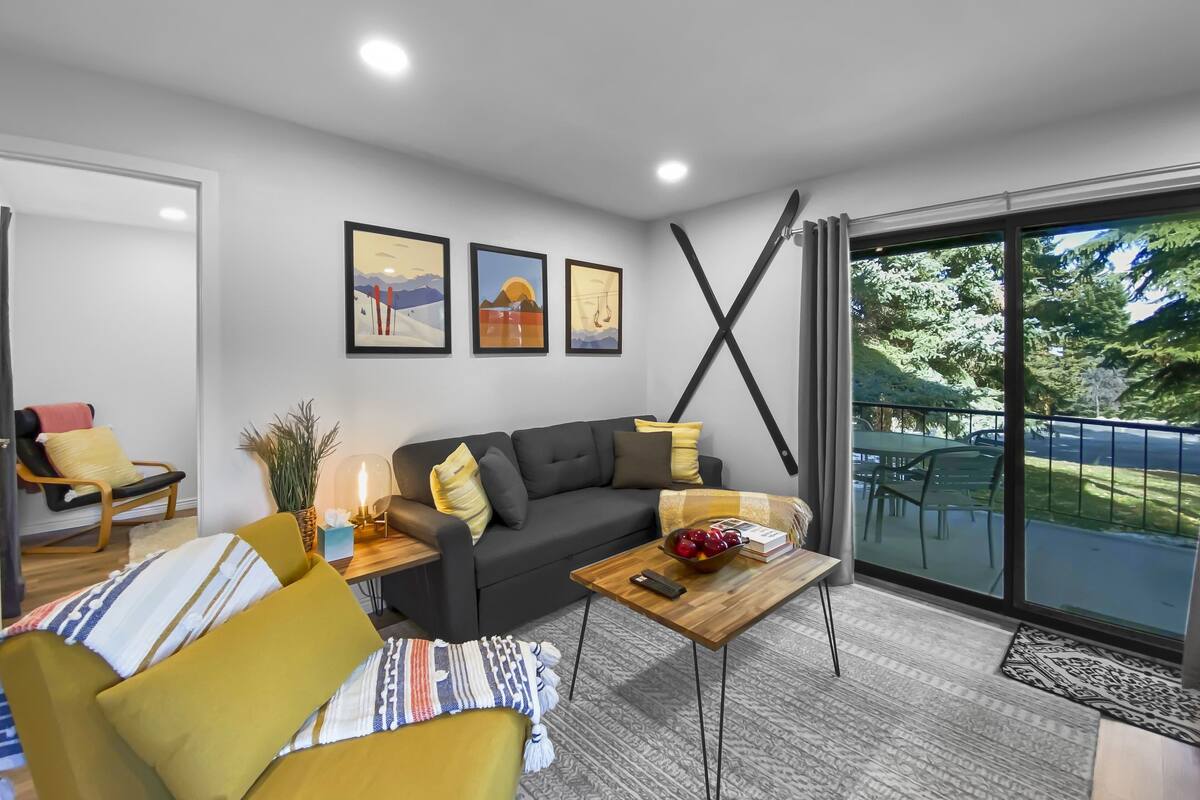 Un salon moderne avec un canapé, une table basse et un fauteuil. Trois tableaux encadrés ornent le mur, ainsi qu’une plante et une fenêtre donnant sur un paysage verdoyant.