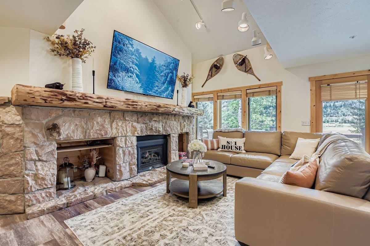 A cozy living room with a stone fireplace, a large flat-screen TV, a leather sofa, and a view of the outdoors through large windows.
